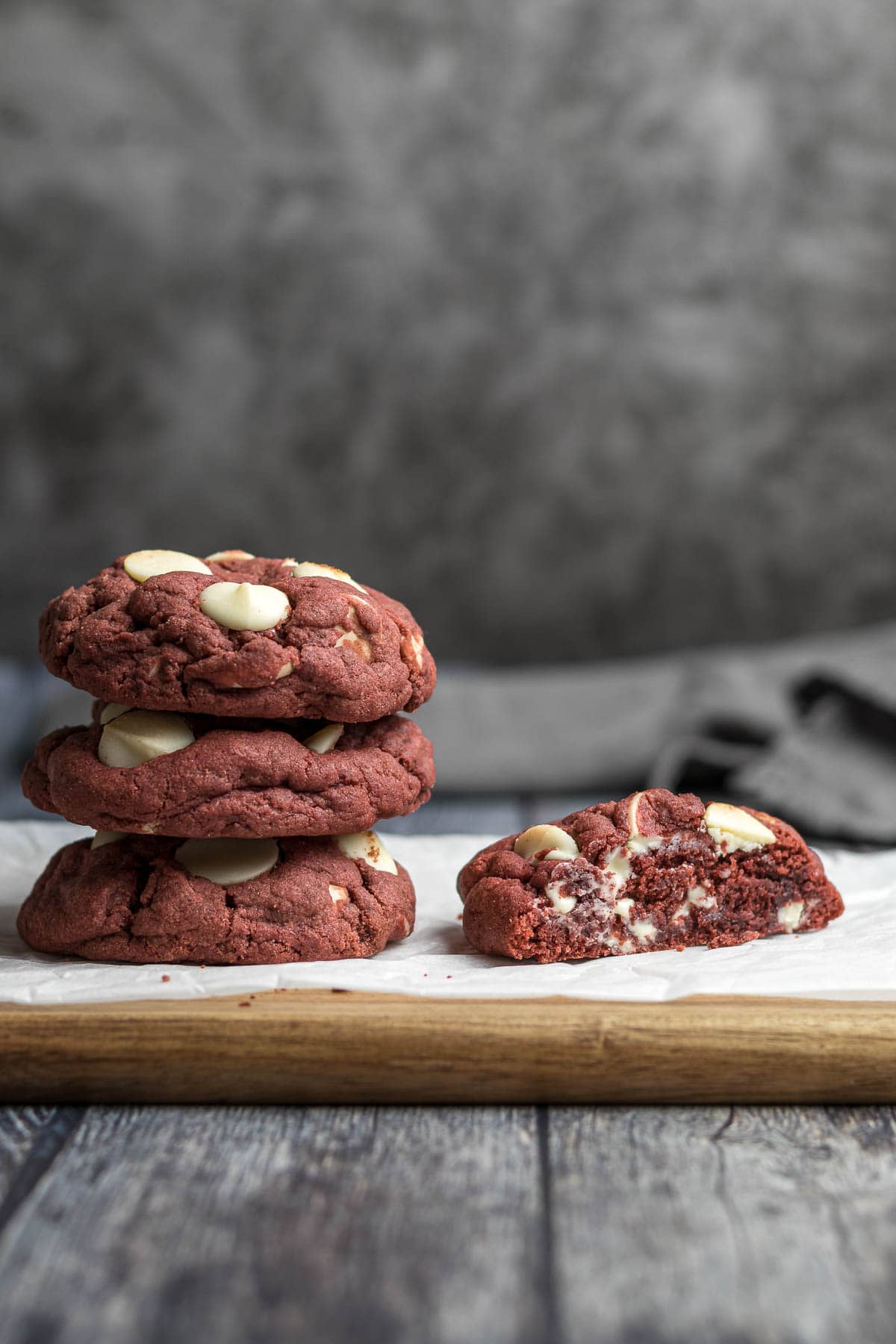 Three red velvet cookies stacked on each other next to a red velvet cookie sliced in half to show the cross section and white chocolate chips