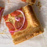 Sliced tomato puff pastry tart showing cheese, tomatoes and red onion.