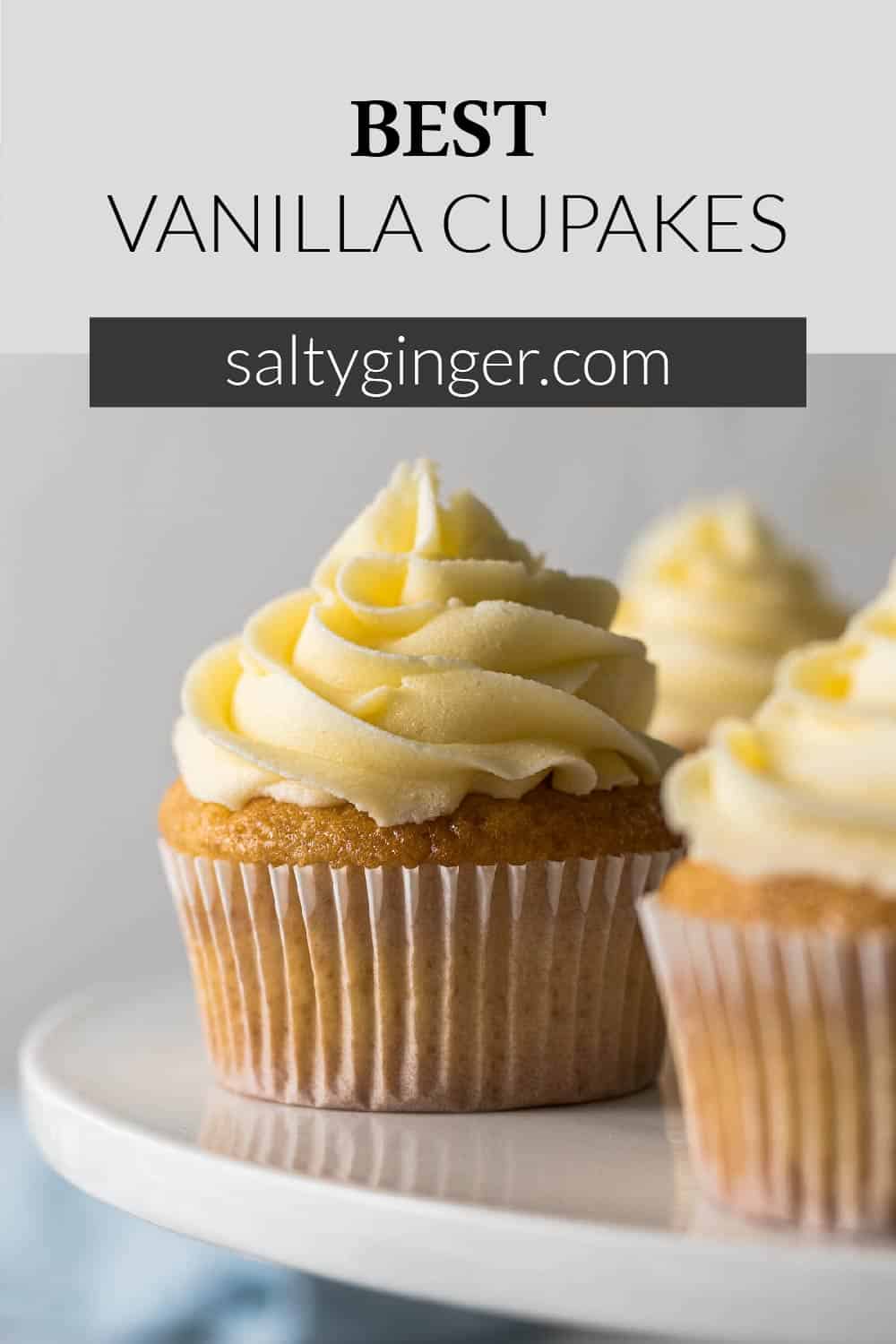 Pin - Vanilla cupcake with a large swirl of vanilla buttercream frosting on a cake stand.