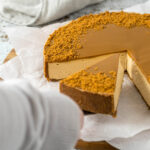 A hand picking up a slice of cheesecake with a cake server.