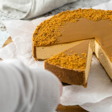 A hand picking up a slice of cheesecake with a cake server.
