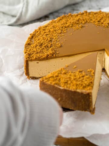 A hand picking up a slice of cheesecake with a cake server.